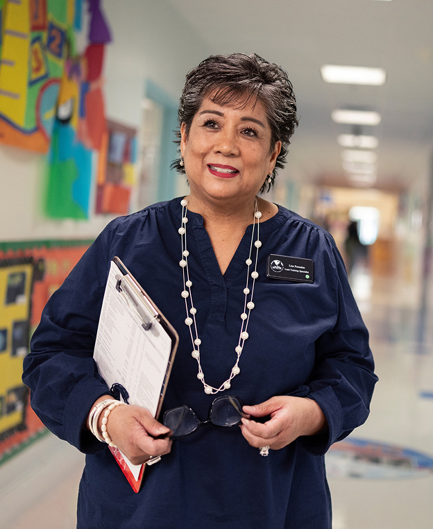 A smiling woman holding a clipboard and a pair of glasses in an elementary school hallway