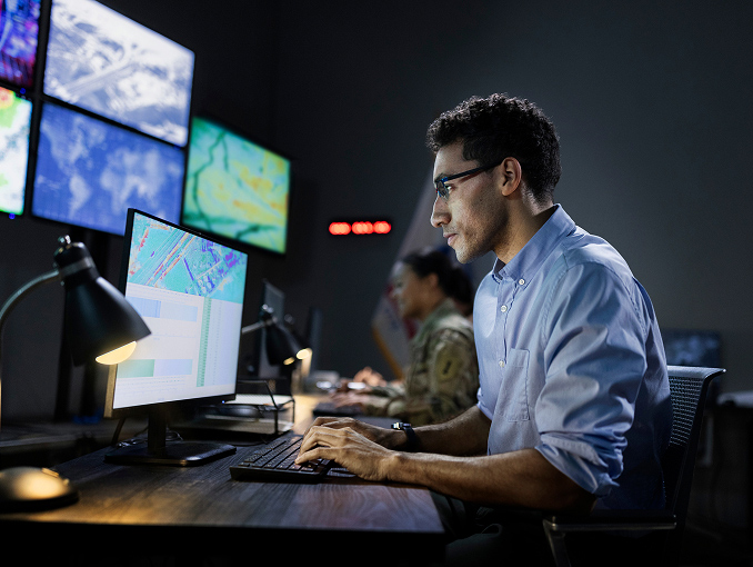 A male IT specialist working at a desktop computer with multiple large monitors on the wall in front of him