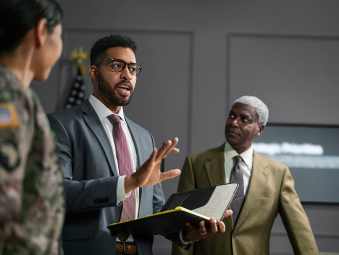 A man in a suit and glasses holding a notebook and leading a meeting comprised of civilian and military personnel