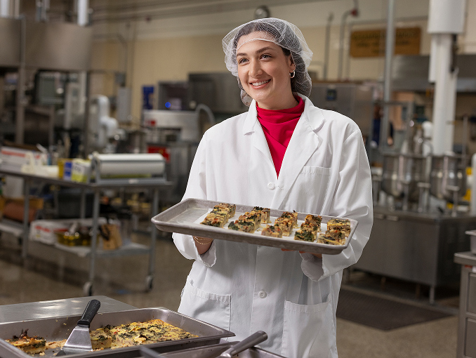 A female food tech wearing a lab coat and hairnet holding a tray of food samples